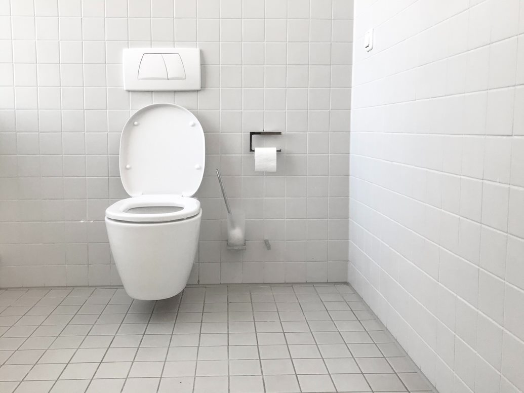 White tiled bathroom stall with toilet, toilet paper holder, toilet brush, and seat cover dispenser above bowl