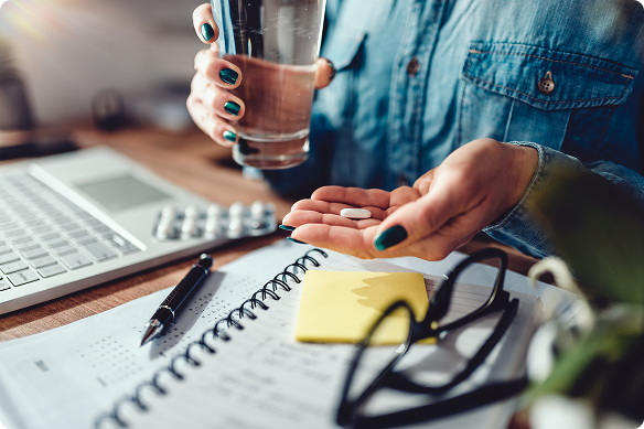 A woman’s hands holding a water glass in one hand and a pill in another, surrounded by a laptop, pen, and paper.