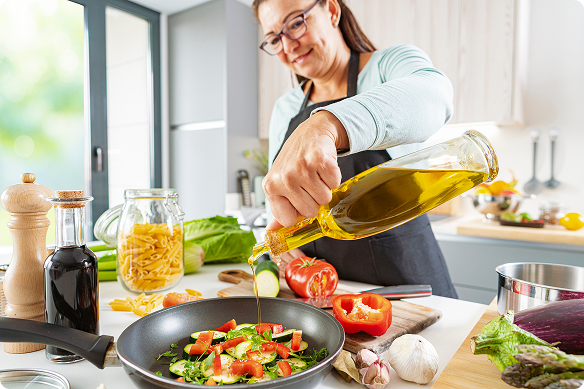 A woman pours olive oil into a pan of tomatoes, zucchini, and herbs on the stovetop in her kitchen.
