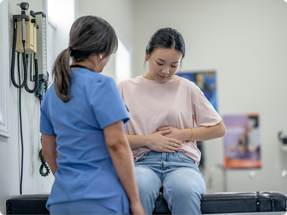 A woman looks down to her stomach and holds it with her hands while discussing symptoms with a nurse.