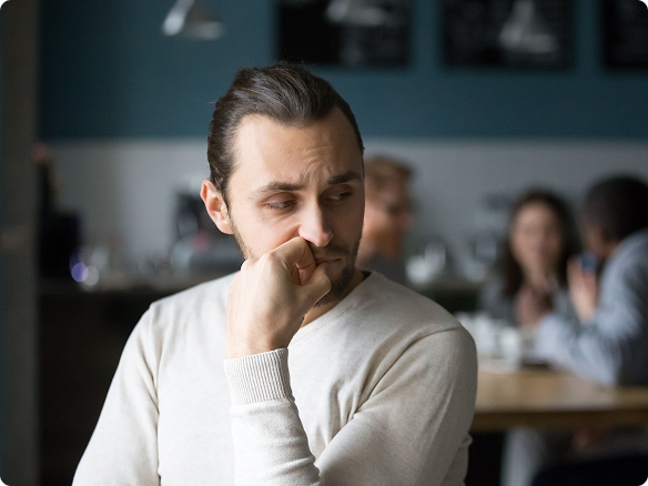 Pensive man in a light sweater rests his chin on his hand in a busy café, looking worried and distracted as blurred patrons chat behind him.
