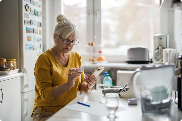 An older woman looks for information on her medication on her phone while she sits in her kitchen.