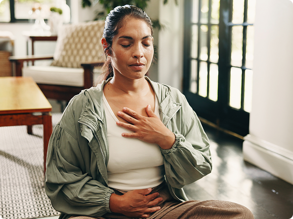 Woman sitting in her living room, hands on heart and stomach, meditating