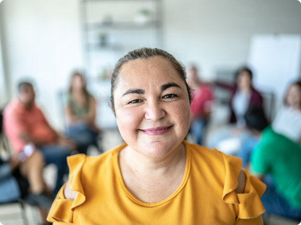 Woman in yellow shirt smiles at camera with blurred support group behind her.