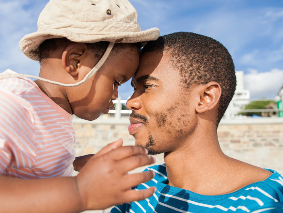 A father and young child touch foreheads affectionately outdoors; the child wears a sun hat, sharing a warm, loving moment.