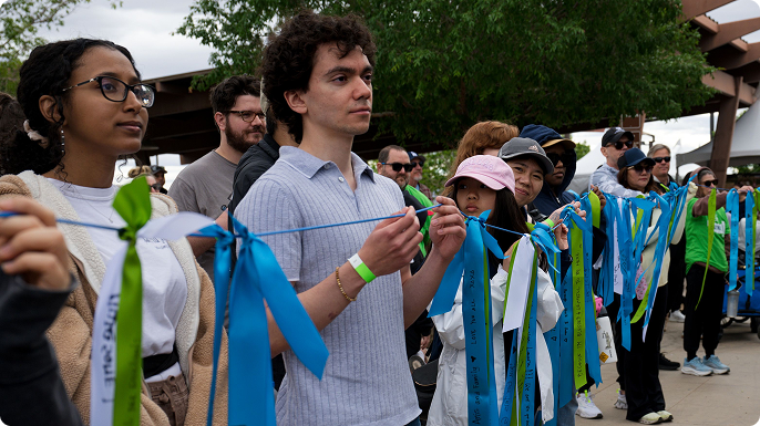 Volunteers hold colorful blue and green ribbons outdoors, focused and hopeful, while a diverse crowd gathers behind them at a Take Steps event.