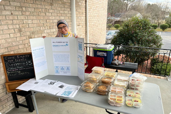 Smiling volunteer displays a fundraiser sign and brochure beside tables of packaged sandwiches, snacks, and desserts on a porch.