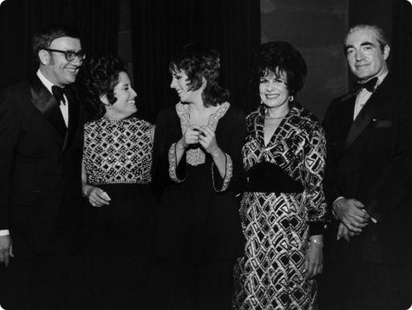 Black-and-white photo of five formally dressed adults at an event, smiling and chatting together in a relaxed, celebratory atmosphere.