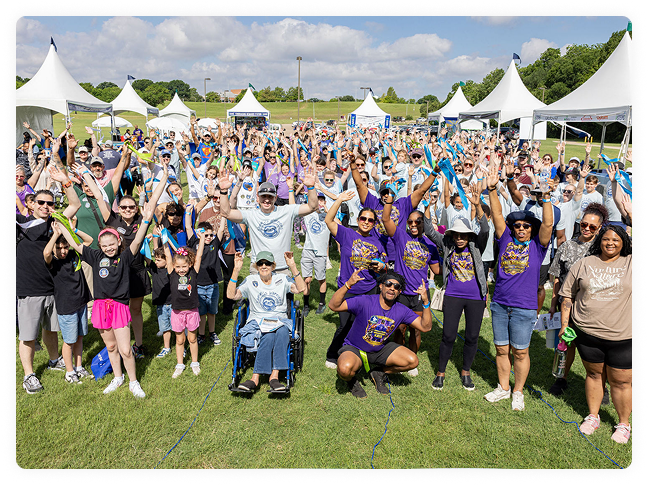 Large, diverse crowd cheers outdoors beneath white event tents, wearing colorful shirts and raising hands in a joyful, inclusive celebration at a Take Steps event.