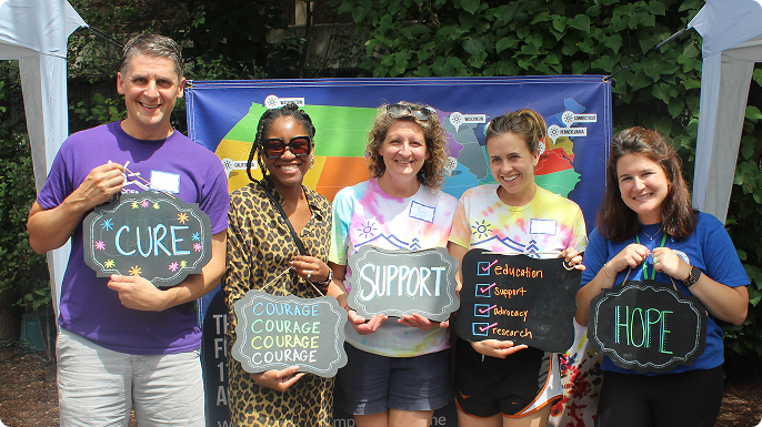 Five smiling adults pose outdoors with colorful banners, holding signs reading “CURE,” “COURAGE,” “SUPPORT,” and “HOPE” at a Take Steps event.