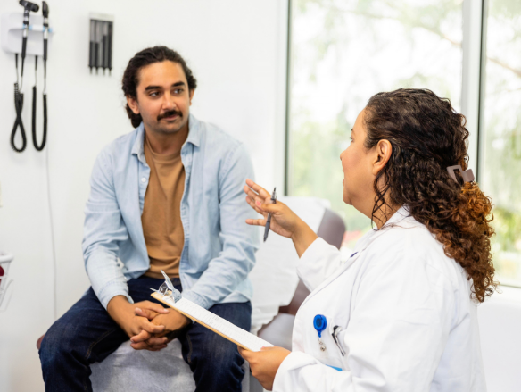Doctor in a white coat discusses care with a seated male patient, holding a clipboard in a bright exam room; both appear calm and attentive.