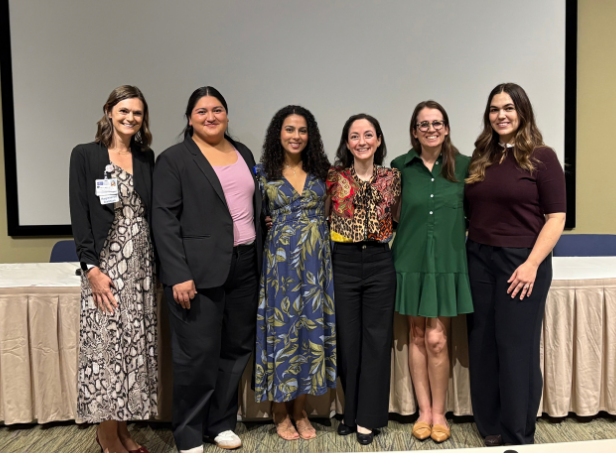 Six women stand arm in arm at an event, dressed in professional attire before a blank screen and table, smiling warmly for a group photo.