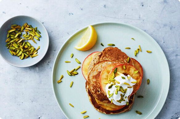 Overhead view of golden pancakes topped with yogurt, peach slices, and pistachios on a pale plate, with lemon wedge and extra nuts nearby.