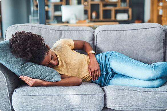 A tired Black woman sleeps curled up on a gray sofa, resting her head on a blue pillow in a living room.