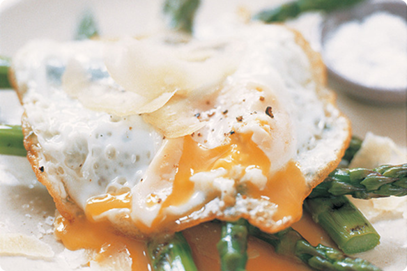 Close-up of a runny fried egg with golden yolk over green asparagus, creating a warm, appetizing brunch scene.