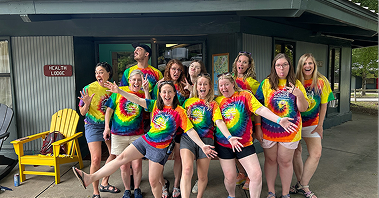 Smiling group of Camp Oasis campers in colorful tie-dye shirts pose playfully on a cabin porch, with outstretched arms and bright, cheerful energy.