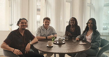 Four diverse IBD patients smile around a round café table, enjoying coffee and conversation in a bright, welcoming room with tall windows.