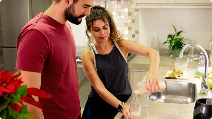 Standing in the kitchen, a woman pours a glass of kefir for her male friend