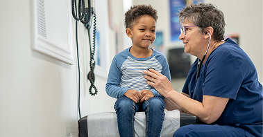 Young boy sits on an exam table as a healthcare provider in scrubs uses a stethoscope during a pediatric gastroenterology checkup