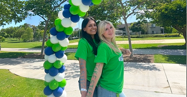 Simran Vaswani and friend in green shirts pose beneath a blue, green, and white balloon arch outdoors on a sunny day at a Take Steps walk