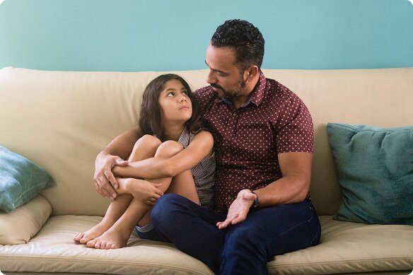 A man sits with his arm around his young daughter while conversing