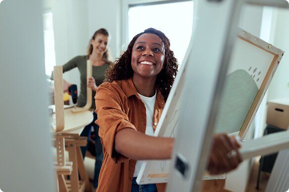 A young adult woman smiles as she holds a painting and grips a ladder, getting ready to hang it on her wall