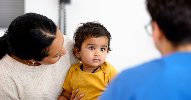 A mother holds her toddler while conversing with a doctor