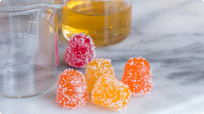 Two beakers sit on a marble counter next to five cannabis gummies