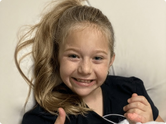 Image of a young girl smiling at the camera and giving a ‘thumbs up”