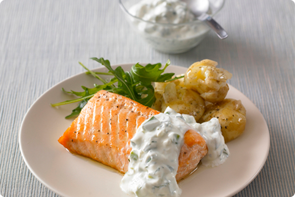 Plate of grilled salmon with creamy herb sauce, roasted potatoes, and greens on a beige dish; a bowl of sauce sits behind.