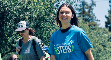 Smiling woman in a blue “Take Steps” charity T-shirt walks on a sunny trail with a woman in a sun hat, surrounded by leafy green trees.