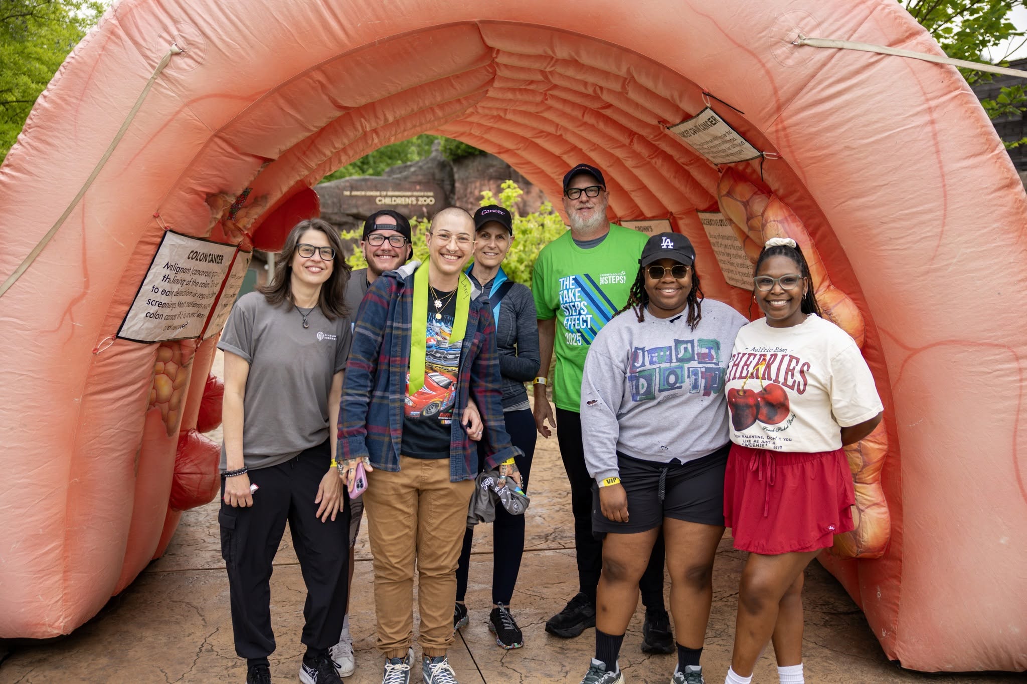 A diverse group of seven smiling people poses inside a pink inflatable tunnel at an outdoor event, creating a cheerful, welcoming atmosphere.