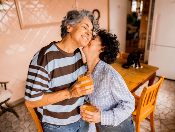 Smiling same-sex couple shares an affectionate kiss in a sunny kitchen, holding orange drinks; a black cat rests on the table behind them.