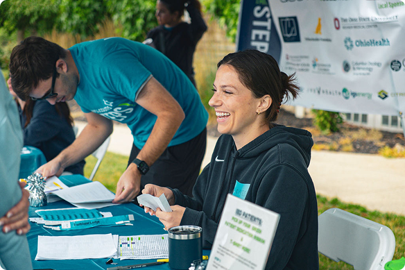 Smiling woman in a black hoodie holds event materials at an outdoor registration table, while a man in teal organizes papers behind her.