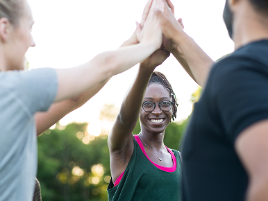 Three diverse teammates celebrate with a high five outdoors; a smiling woman in glasses stands between two blurred teammates, conveying teamwork and joy.