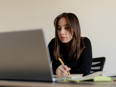 Thoughtful woman looking at her computer and taking notes with pad and pen.