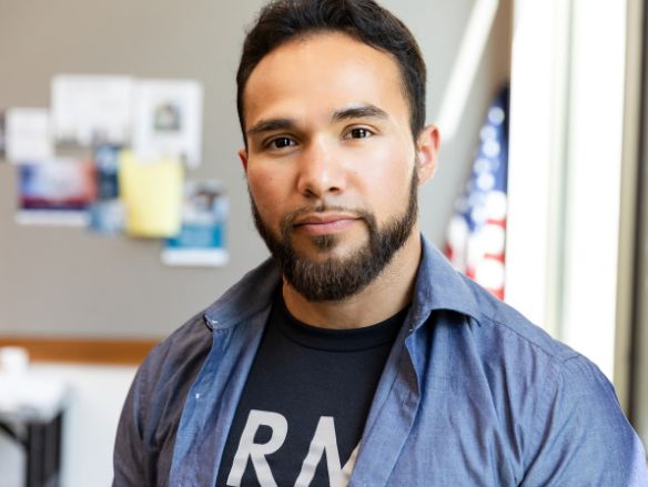 Bearded man in a blue shirt and black “ARMY” T-shirt smiles calmly at the camera in a bright healthcare office.