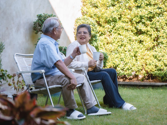 Two older adults sit in garden chairs, smiling and chatting over coffee outdoors, creating a warm, relaxed, companionable mood on a sunny afternoon.