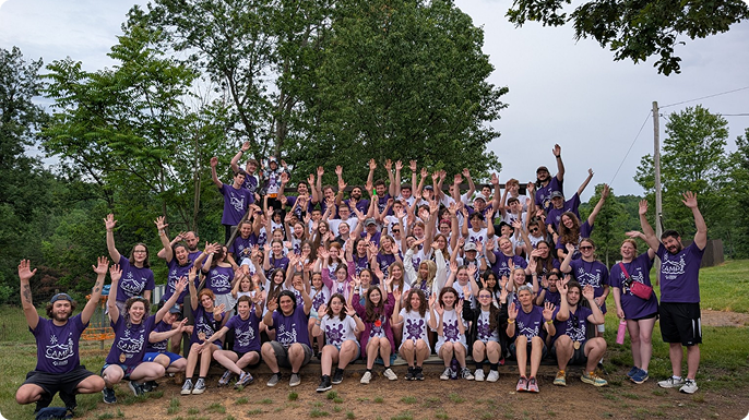Large, diverse group of Camp Oasis campers and counselors in purple shirts poses outdoors, raising hands and smiling together in a joyful, celebratory team photo.