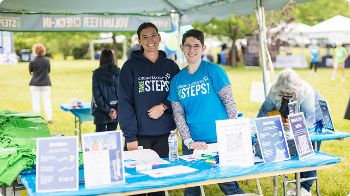 Two smiling women in “Take Steps” shirts stand at a charity information booth outdoors, surrounded by brochures and banners in a sunny park.