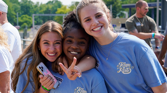 Three smiling young women pose outdoors at an event in blue Team Rio shirts; one holds a phone while another flashes a peace sign.