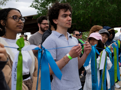 A young man in a blue shirt holds a teal ribbon stretched between his hands at an outdoor event, surrounded by people wearing ribbons and lanyards.