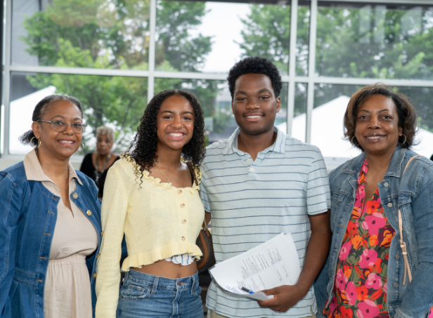 Four smiling family members pose in a bright, windowed lobby; a young man holds paperwork between two older women and a young woman. The mood is warm and welcoming.