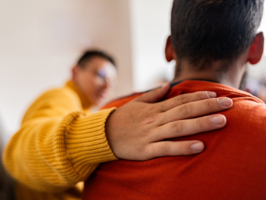A person in a mustard-yellow sweater rests a hand on another person’s red-shirted shoulder, conveying support and comfort in a softly blurred indoor setting.