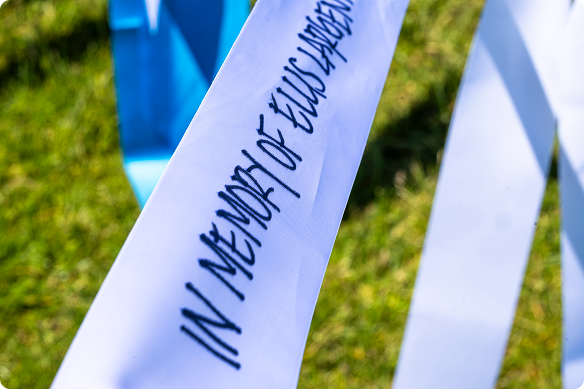 Close-up of white memorial ribbons with handwritten names, including “In memory of...,” against green grass and blue accents, conveying remembrance.
