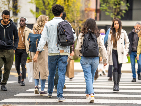 Diverse pedestrians cross a city street at a zebra crossing, carrying backpacks and bags; a busy, casual urban commute feels energetic and everyday.