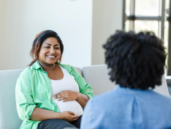 A smiling pregnant woman in a green shirt rests a hand on her belly while speaking with a healthcare provider in a bright, calm consultation room.