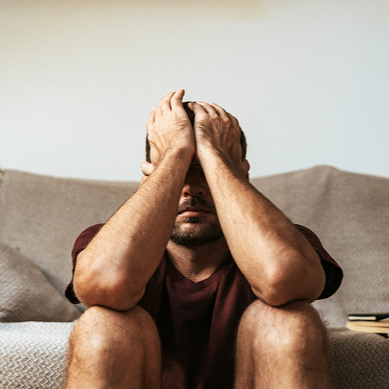 Stressed man sits on a couch with his head in his hands, conveying worry, frustration, and emotional exhaustion in a quiet home.