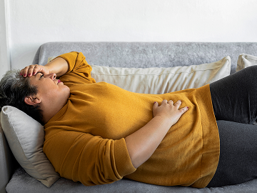 Woman on her couch in a yellow top and grey sweatpants, one hand on her head, one on her stomach, in pain.