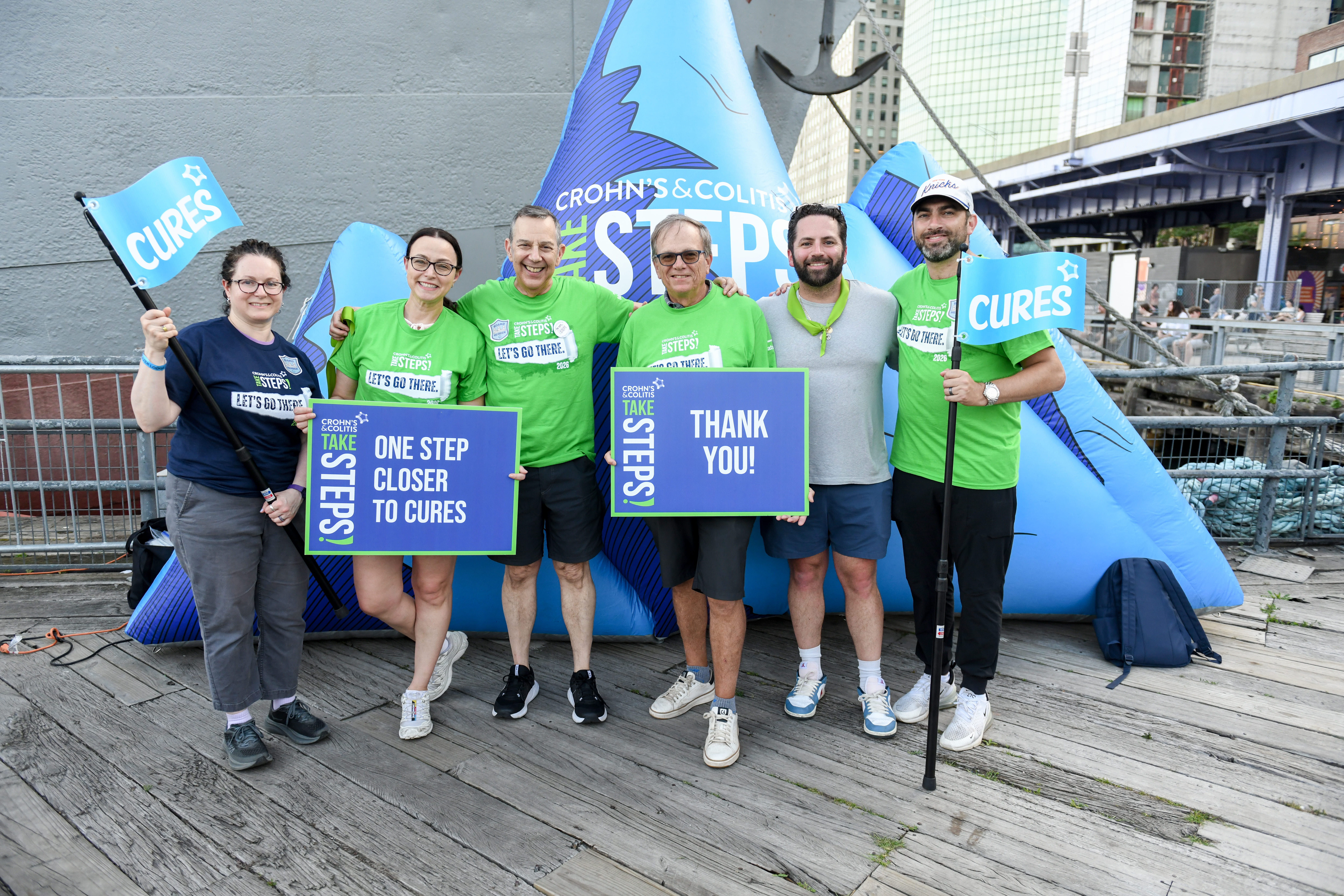 Six smiling charity runners pose before a blue Crohn’s & Colitis Foundation inflatable, holding “Cures,” “One Step Closer to Cures,” and “Thank You” signs.